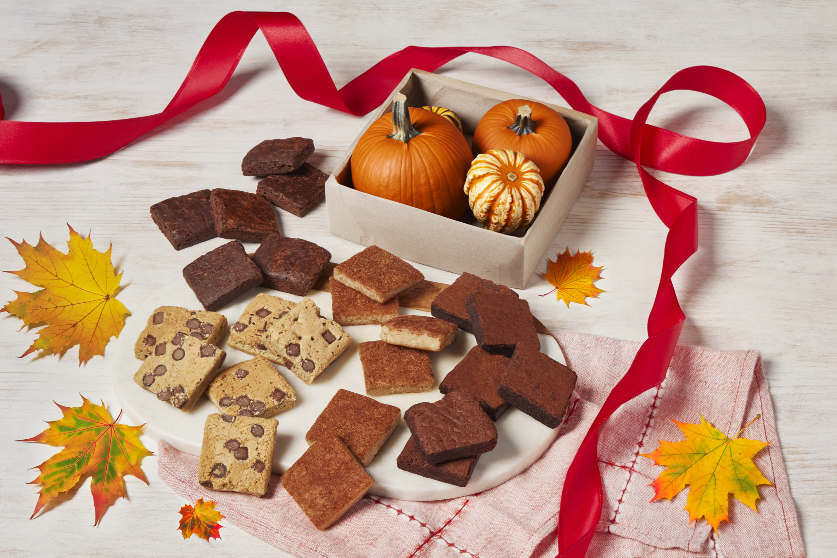 Assorted cookies, small pumpkins, and autumn leaves on a wooden surface with a red ribbon.